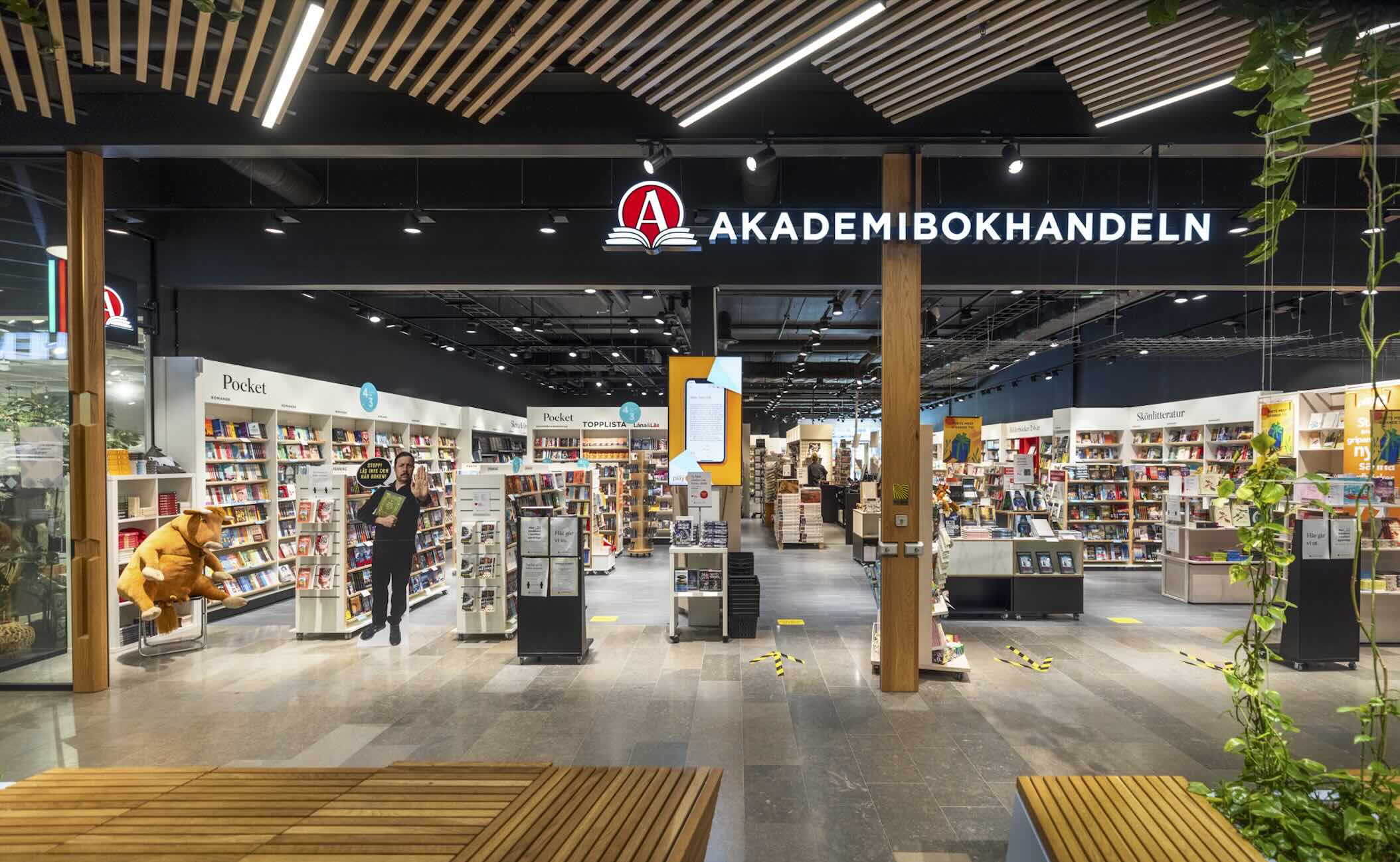 En bokhandel inomhus; en man står i en gång och håller upp en bok, omgiven av hyllor med Pocket och skönlitteratur, samt bänkar och växter i entrén. Text i bilden: AKADEMIBOKHANDELN Pocket TOPPLISTA Skönlitteratur