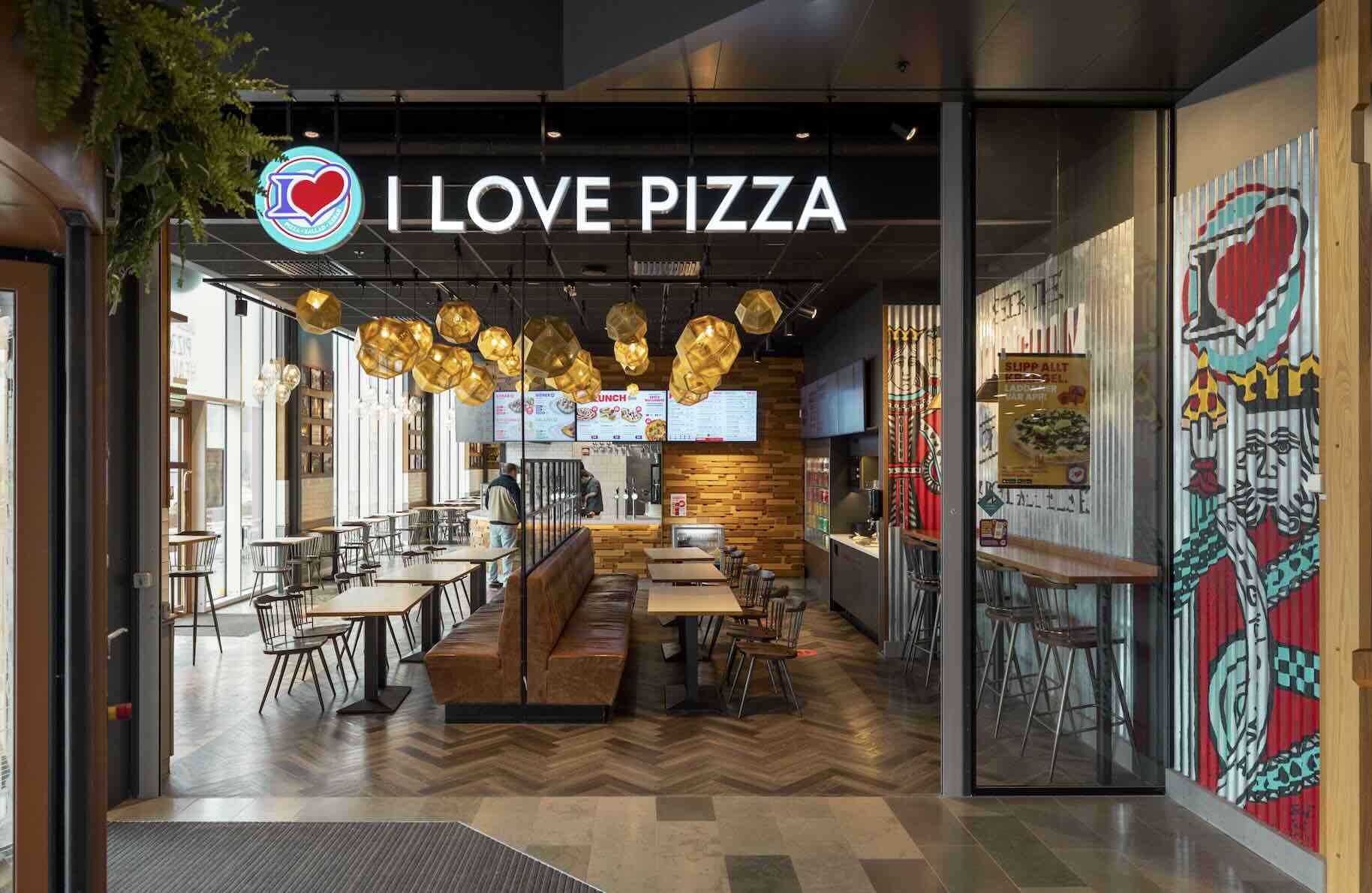 "Restaurangen I LOVE PIZZA i en modern food court, med stor upplyst skylt och hjärtelogotyp. Inuti finns lädersoffor, träbord, flera småbord och hängande gula lampor. Text i bilden: I LOVE PIZZA.