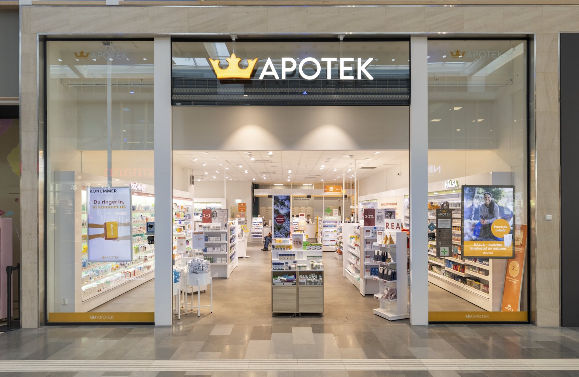 "Öppet apotek i ett shoppingcenter med glasdörrar och guldkrön. Inuti står hyllor med läkemedel och hälsoprodukter; kunder och personal rör sig bland kassor och skyltar. Transkription av text i bilden: - APOTEK - APOTEK - Du ringer in, vi kommer ut - Prova du också!