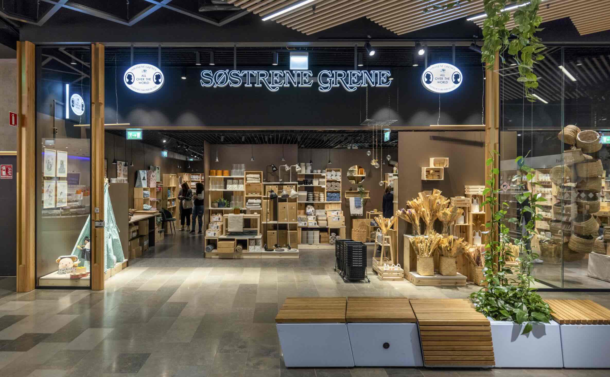 Butikslokal märkt SØSTRENE GRENE, med cirklar som säger ALL OVER THE WORLD, trähyllor fyllda med lådor och prylar. Två kunder bläddrar bland produkterna medan växter står vid ingången.
