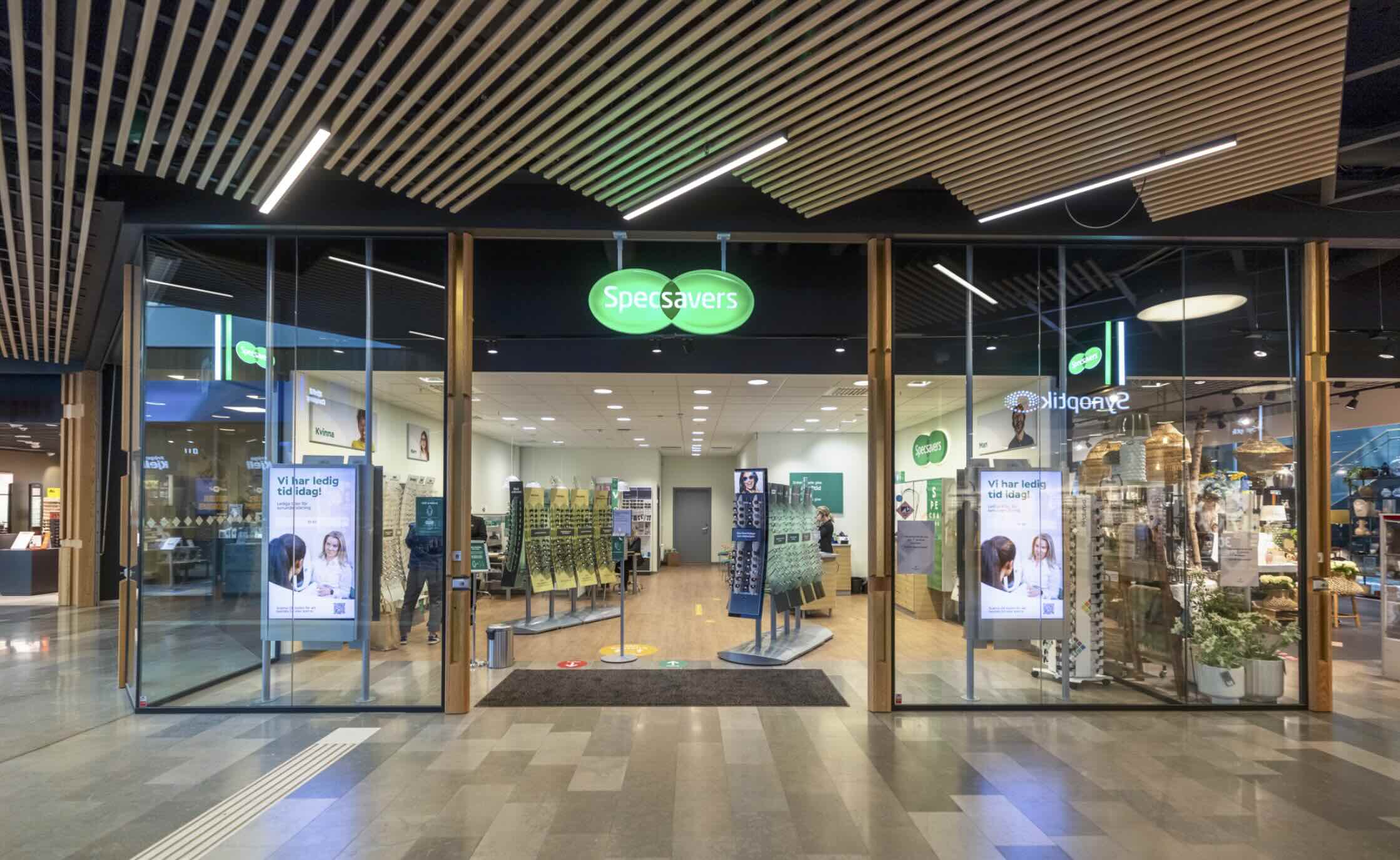 Specsavers-logotyp i två gröna sammanflätade cirklar över ingången till glasfasaden. Inne står glasögonställ och personal vid kassan medan kunder tittar på ramar. Skyltar säger: "Vi har ledig tid idag!"