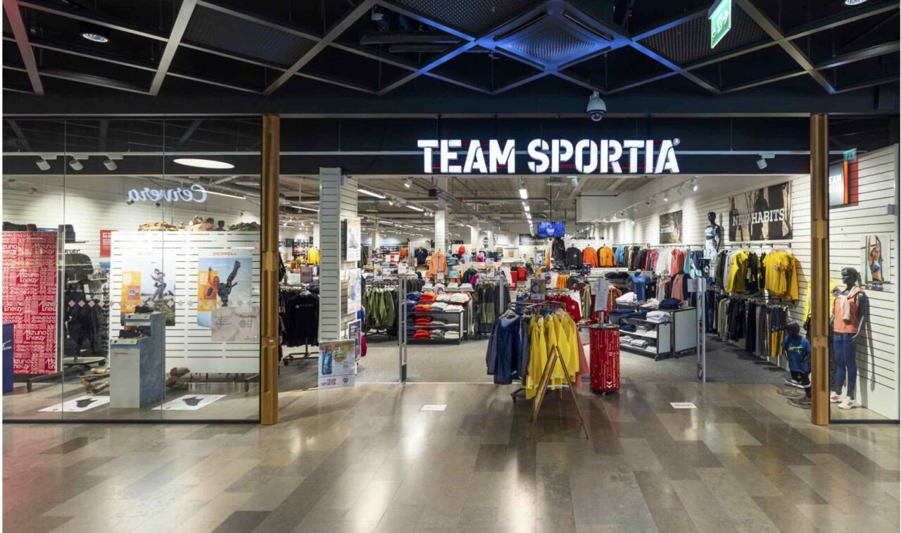 En sportbutik vid gallerian med upplyst skylt TEAM SPORTIA. Exponering av färgade jackor, tröjor och byxor i rader och på bord, medan väggarna visar annonser som MERRELL och NEW HABITS.