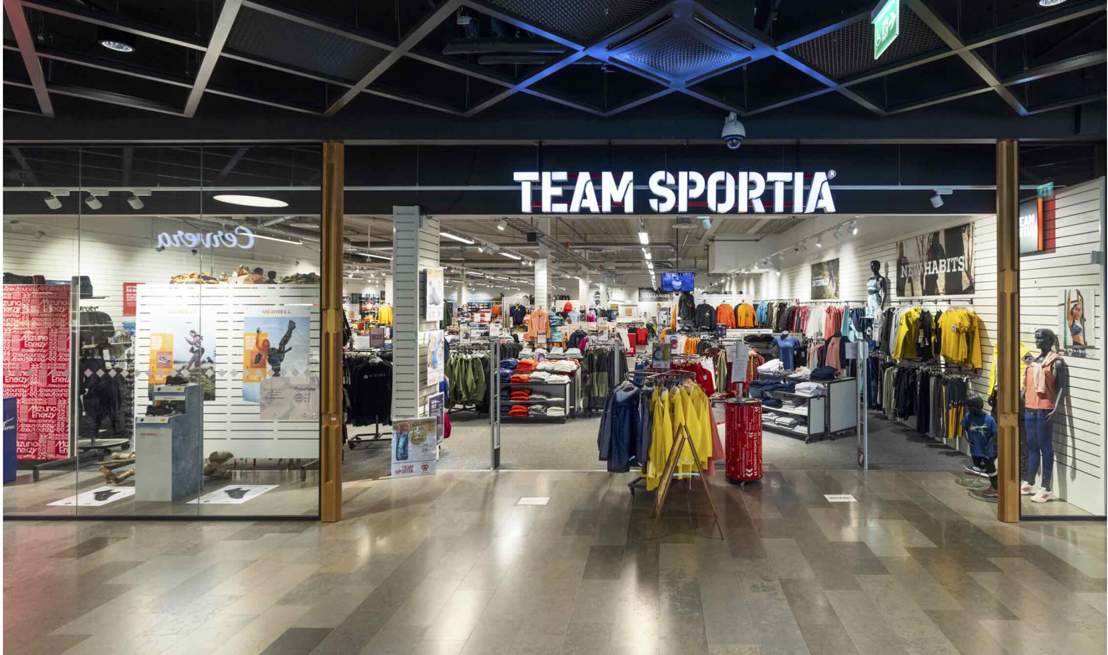 En sportbutik vid gallerian med upplyst skylt TEAM SPORTIA. Exponering av färgade jackor, tröjor och byxor i rader och på bord, medan väggarna visar annonser som MERRELL och NEW HABITS.