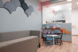 En barnvänlig hörna i ljusblått med en rutmönstrad soffgrupp längs väggen. Ett bord med blå stolar och böcker på vägghylla. En orange djurformad väggdekor och röda nätblommor hänger i taket.