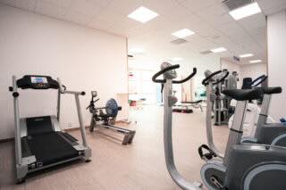 Ett ljust gymrum fullt av träningsredskap: ett löpband står längst till vänster, en roddmaskin mitt i rummet och flera motionscyklar längre in; väggar är ljusa och golvet är trälikt.
