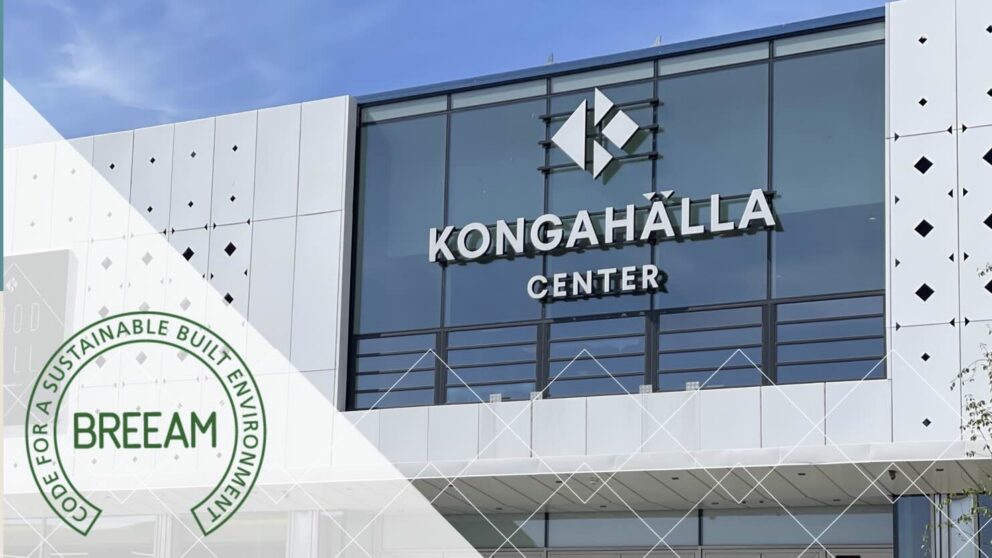 En modern byggnad med glasfasad och vit panel står i bildens centrum. På fasaden läses "KONGAHÄLLA CENTER" under en geometrisk logotyp. Till vänster nedtill syns en grön rund BREEAM-stämpel med ordet "BREEAM" i mitten och runt kanten texten "FOR A SUSTAINABLE BUILT ENVIRONMENT".
