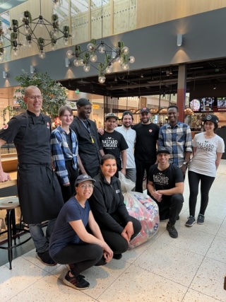 Personal I Food Hall "En grupp personer står tillsammans framför en snabbmatsrestaurang i ett ljust köpcentrum, där flera bär Bastard Burgers-kläder. De ler mot kameran medan ett stort paket pant ligger på marken. BASTARD BURGERS"