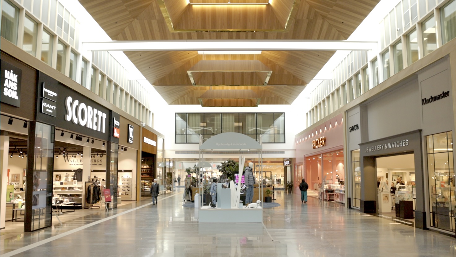 Den centrala utställningen i en ljus inomhusgalleria: en vit, rektangulär monter med kläduppsättningar och mannequiner; omgiven av butiker längs sidorna och ett tak av träpaneler. Text i bilden: SCORETT GANT JEWELLERY & WATCHES SMYCKA Upplev något annorlunda KLOCKMASTER