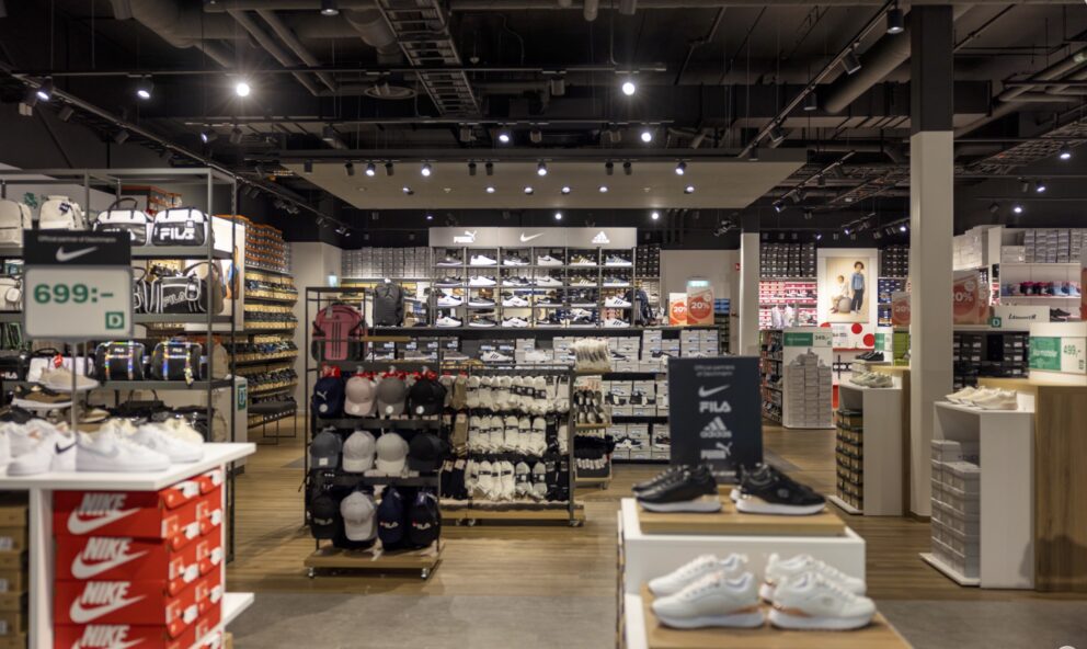 Skohylla visar skor och kepsar i en sportbutik; lampor lyser över displayen medan priset 699:- och 20% rabatt syns. Märken: Nike, FILA, Adidas, Puma.