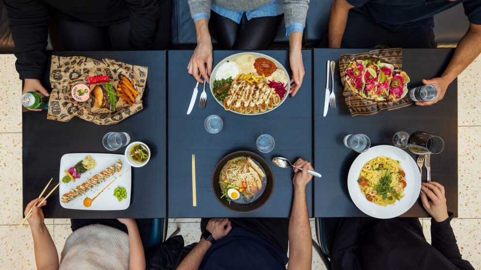 Mitt på bordet ligger en stor tallrik med skivade rullade maträtter och sallad; runtom syns sushi, ramen och pasta samt färgglada tacos i varierande färger.