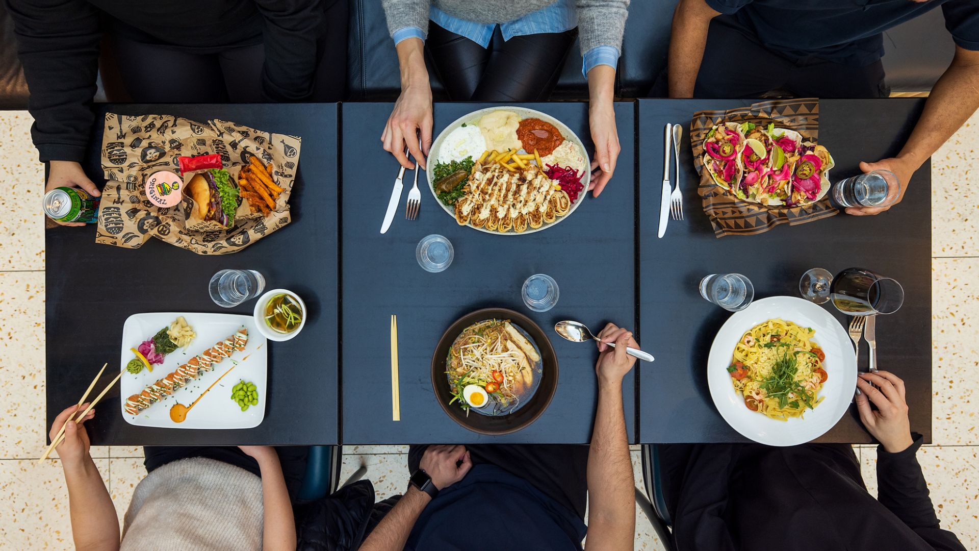 Mitt på bordet ligger en stor tallrik med skivade rullade maträtter och sallad; runtom syns sushi, ramen och pasta samt färgglada tacos i varierande färger.
