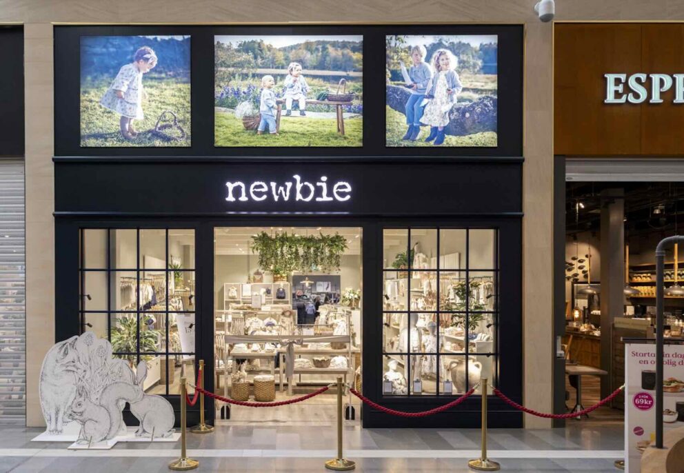Butiksfasad tillhörande nyöppnade butiken 'newbie' med öppna dörrar; inuti syns kläder, växter och lampor. Omgivningen är ett köpcentrum med andra butiker; synligt är även 'instagram'-symbol nära ingången.