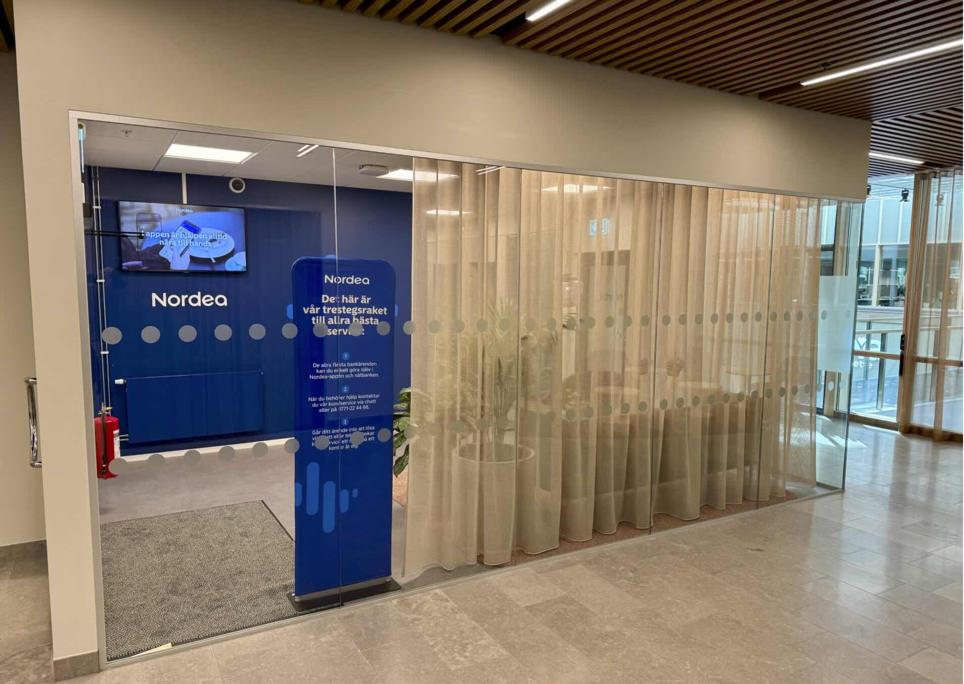 "Nordea-relaterad bankhall genom glasväggar; ett blått informationsstånd i förgrunden; beige gardiner och träpaneler i taket ger en modern bankmiljö. Text i bilden (transkriberad): Nordea Nordea Nordea