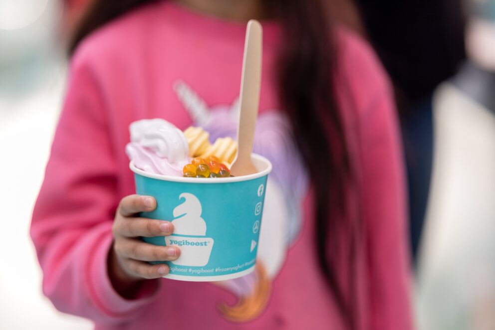Flicka i rosa tröja äter Yogiboost-glass med topping.
