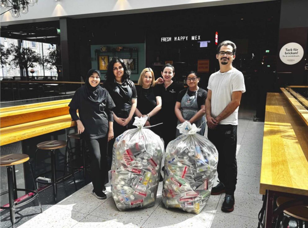 Sex personer står och ler bredvid två soppåsar fyllda med plastflaskor och burkar. Miljön är ett cafékök med träbänkar. Texter: FRESH HAPPY MEX; Tack för brickan! Du är omtänksam.