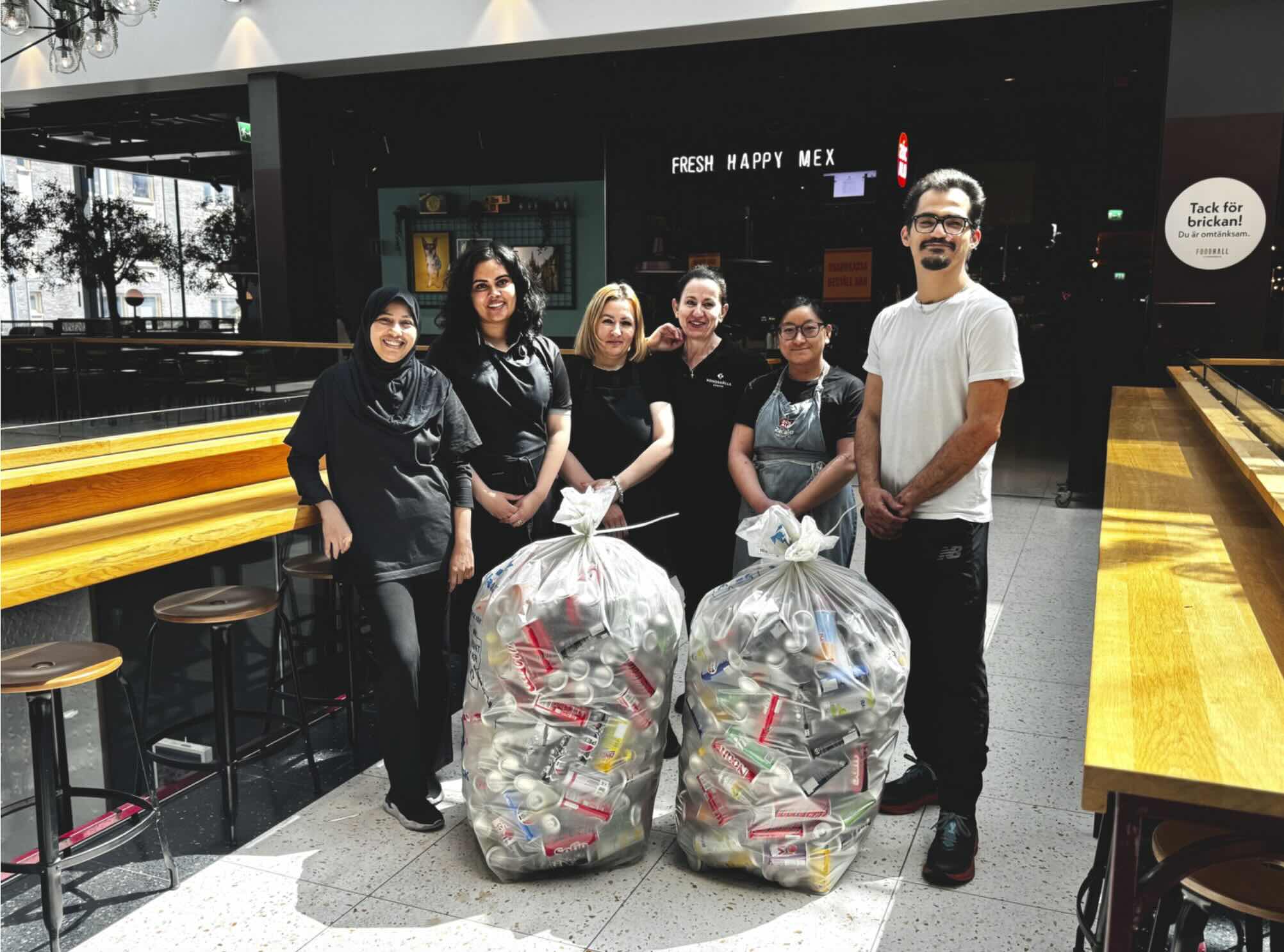 Sex personer står och ler bredvid två soppåsar fyllda med plastflaskor och burkar. Miljön är ett cafékök med träbänkar. Texter: FRESH HAPPY MEX; Tack för brickan! Du är omtänksam.
