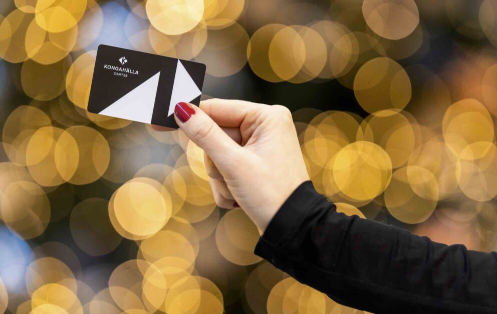 Ett svart kort med vitt geometriskt mönster hålls mellan tummen och pekfingret av en hand med röda naglar; texten KONGAHÄLLA CENTER syns; bakgrunden består av varma gyllene ljus.
