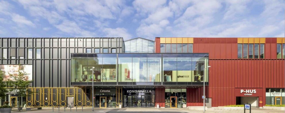 Fasaden till ett modernt köpcentrum domineras av en glasentré i mitten, svarta metallpaneler till vänster och röda/orangea paneler till höger. Text i fasaden: gina tricot, Cervera, KONGAHÄLLA, P-HUS UTFART, Närhälsan.