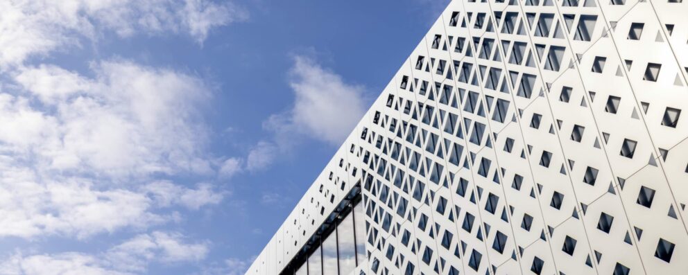 Fasaden av en vit byggnad dominerar bilden och visar ett upprepat diamantformat fönstermönster längs en diagonal yta; bakgrunden är en klar blå himmel med några vita moln.