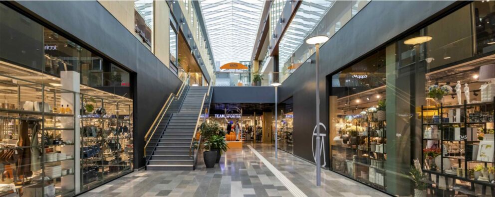 "Mitt i en ljus inomhusgalleria står en bred trappa bredvid en rulltrappa som leder uppåt; glasbutiker, växter och ett genomskinligt tak omger scenen. Text i bilden: TEAM SPORTIA IEMS