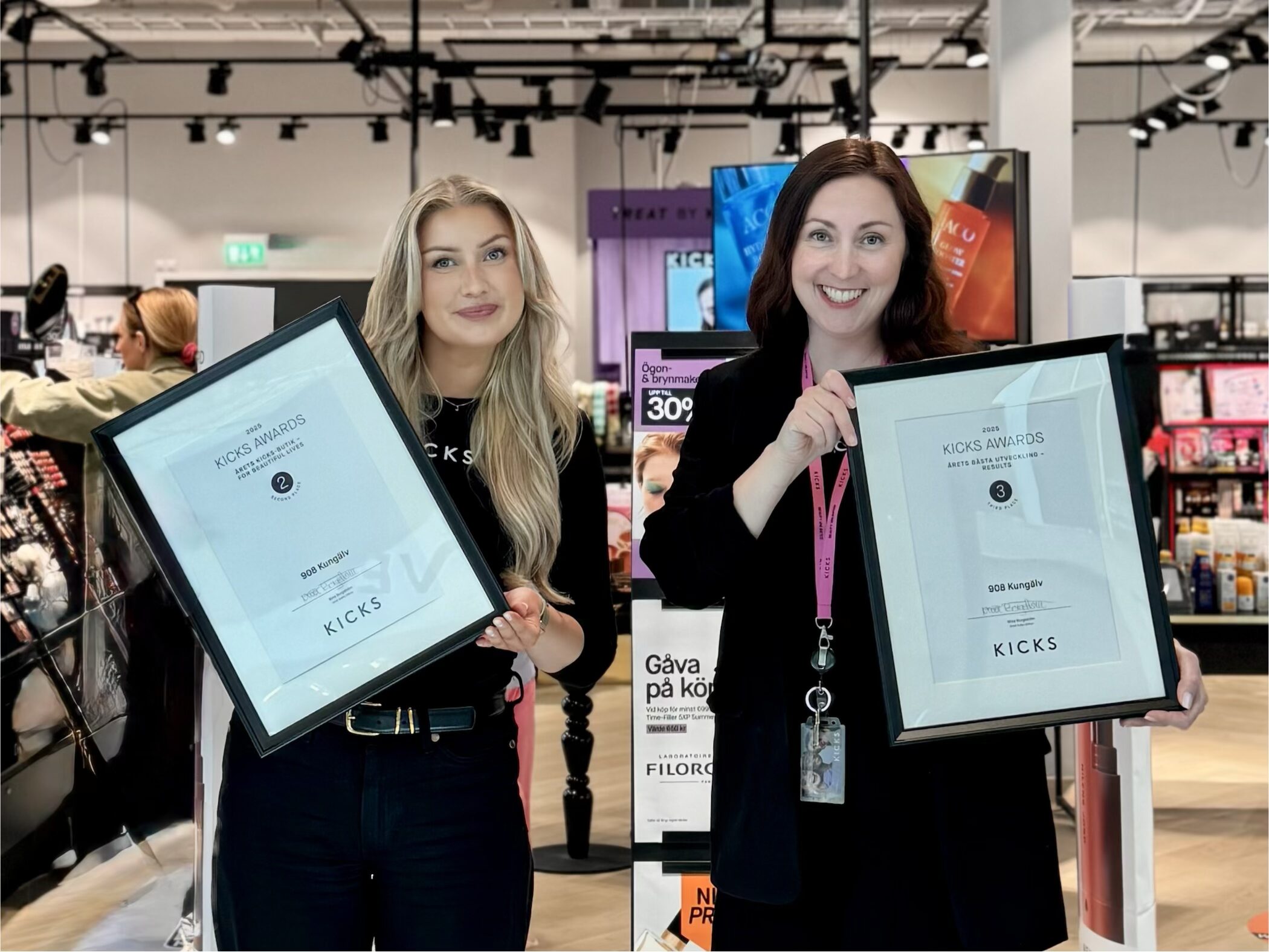 Två kvinnor håller upp diplom, ståendes framför Kicks butik inne i centret
